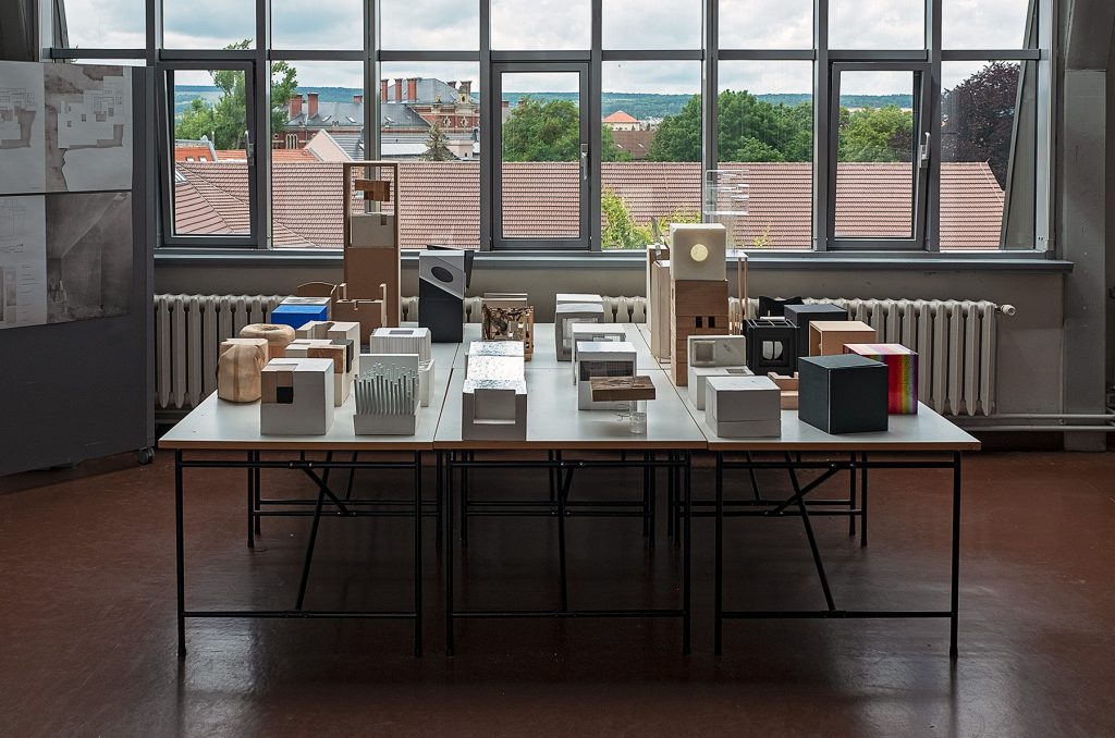 Ein Tisch mit vielen unterschiedlich gestalteten kubischen Formen vor einem großen Atelierfenster. Mit Link zum Beitrag: Weimar, staatliches Bauhaus.